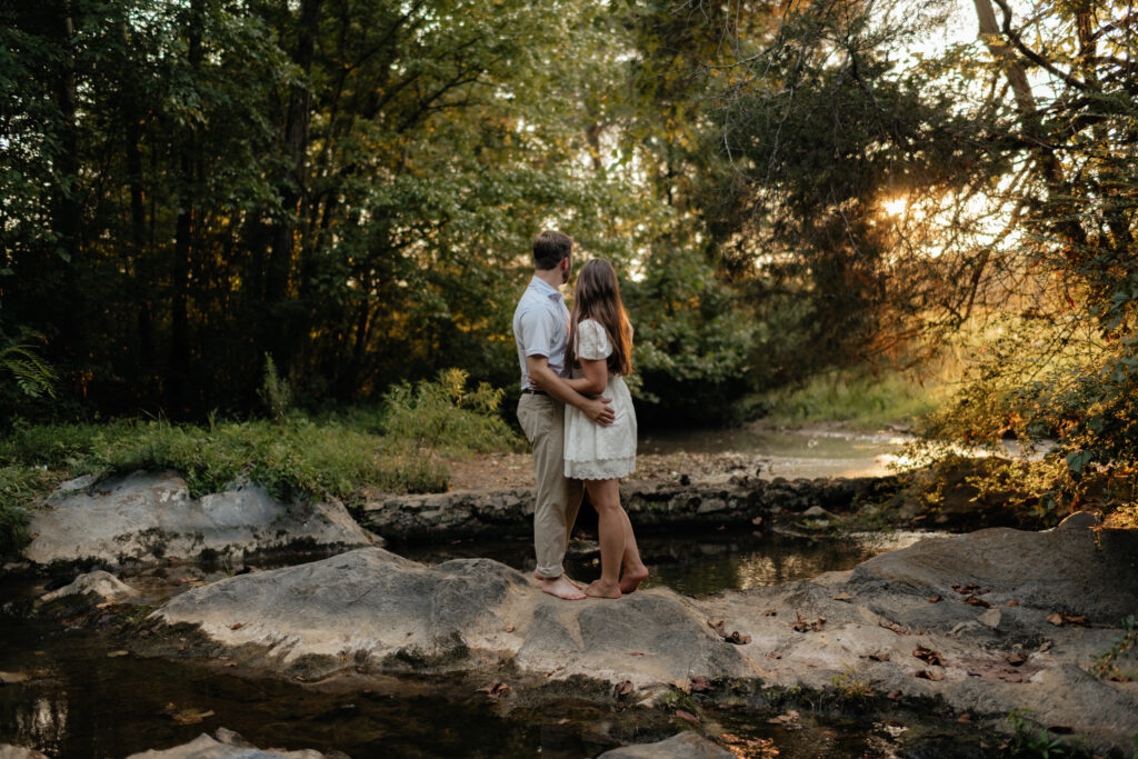 a couple pauses to watch the sun set as they cross a creek during their summer field engagement photos in calera, alabama, photography by Birmingham alabama wedding photographer Sarah Mismash