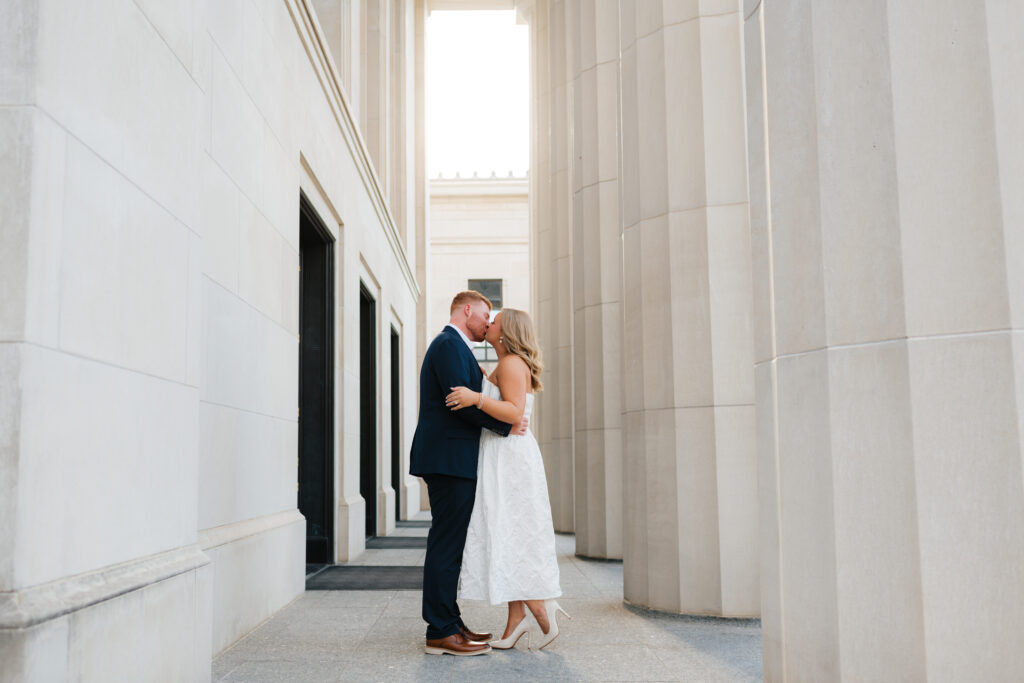 a couple kisses between the columns of the federal building in downtown Tuscaloosa during their engagement photos with alabama wedding photographer Sarah Mismash