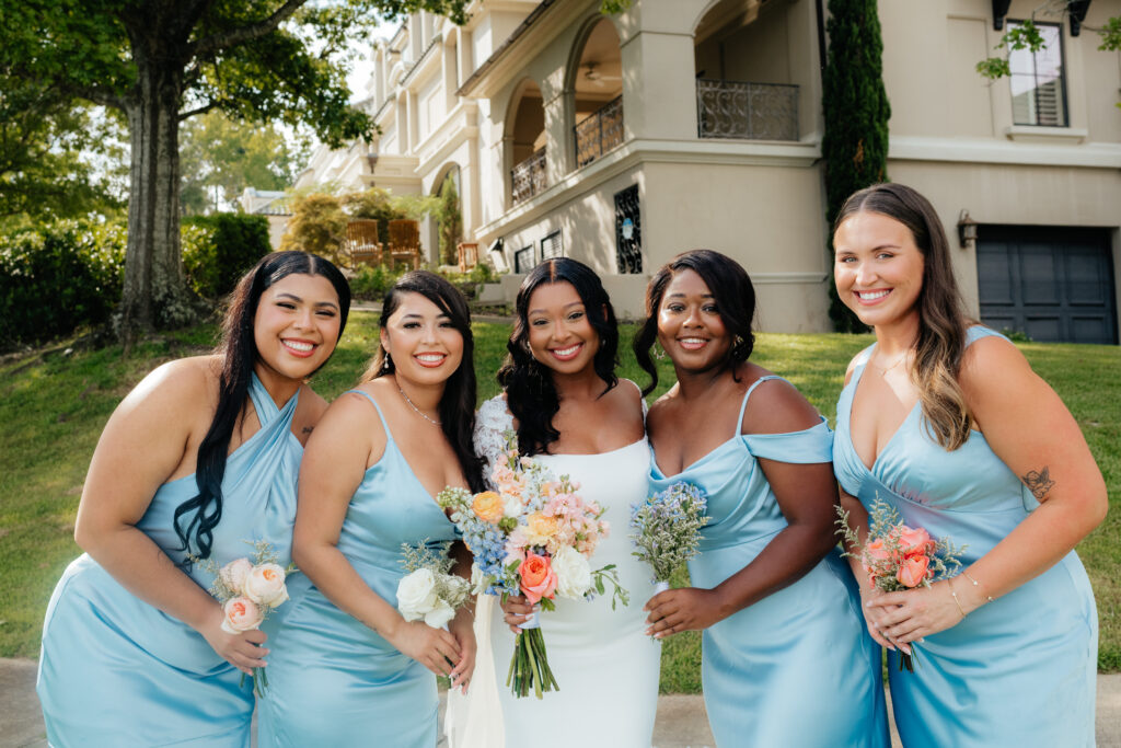 a bride and her bridesmaids dressed in blue satin with pink and white and blue florals smile for alabama wedding photographer Sarah Mismash during a summer wedding at lake Martin near Alexander city