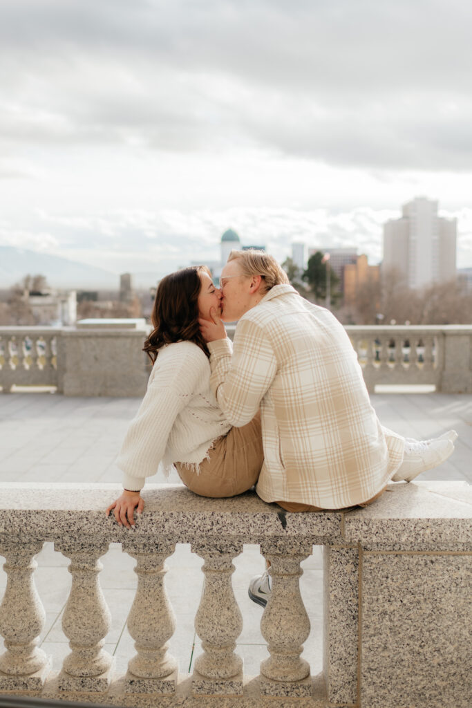 a couple kisses with the salt lake city skyline behind them during their engagement photos at the state capitol with olympia engagement photographer sarah mismash