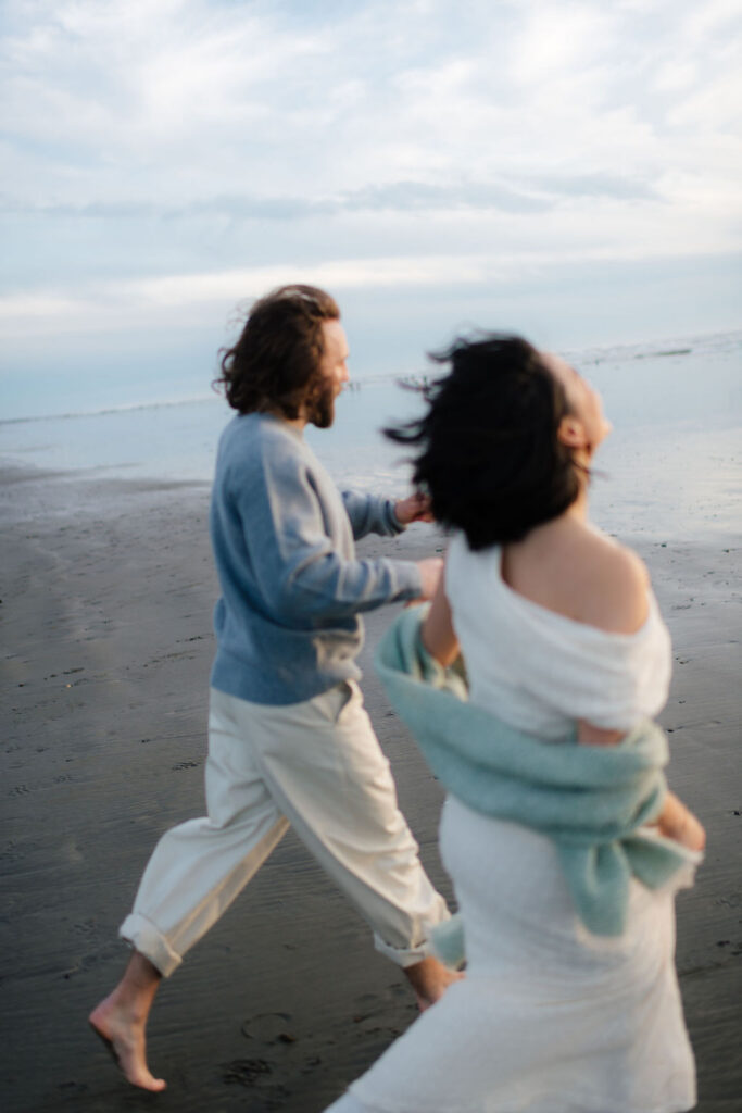 a couple runs through the waves at twin harbors state park during their engagement photos on the washington coast with Westport washington engagement photographer Sarah Mismash