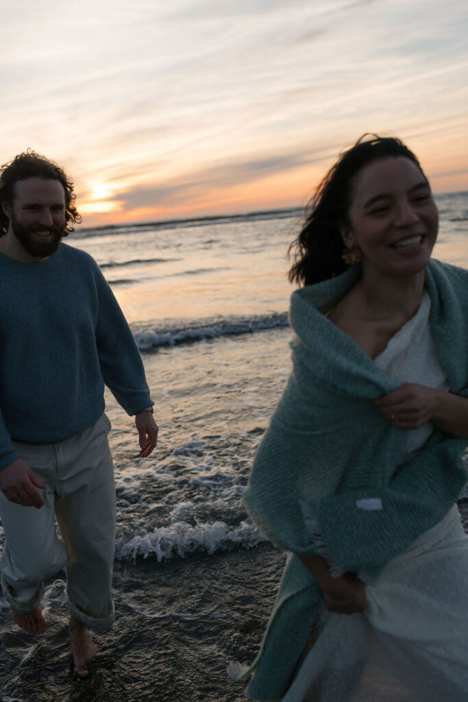 a couple runs through the waves during the sunset at twin harbors state park during their engagement photos on the washington coast with Westport washington engagement photographer Sarah Mismash