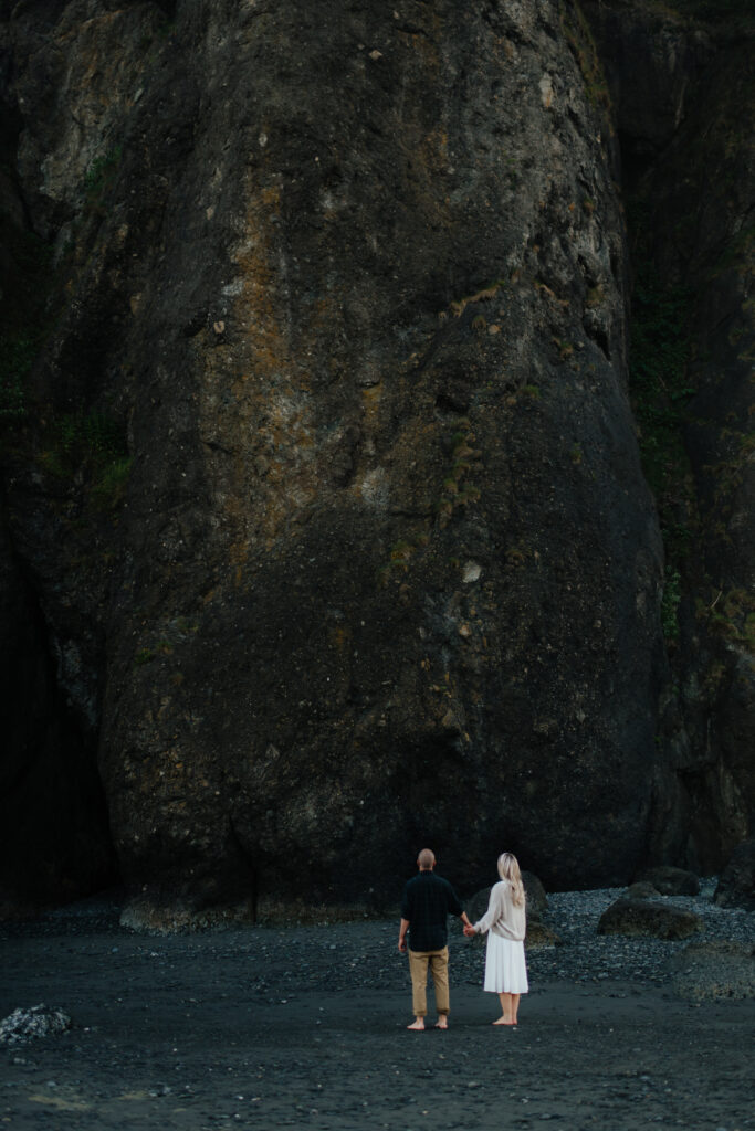 a couple stares up at a massive stone cliff at ruby beach near forks, washington during their summer engagement photos