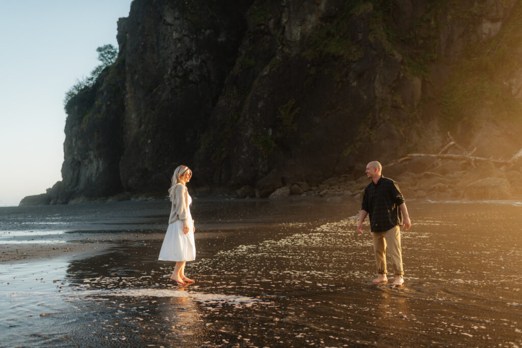 a couple walks across ruby beach during golden hour during their summer engagement photos in forks, washington