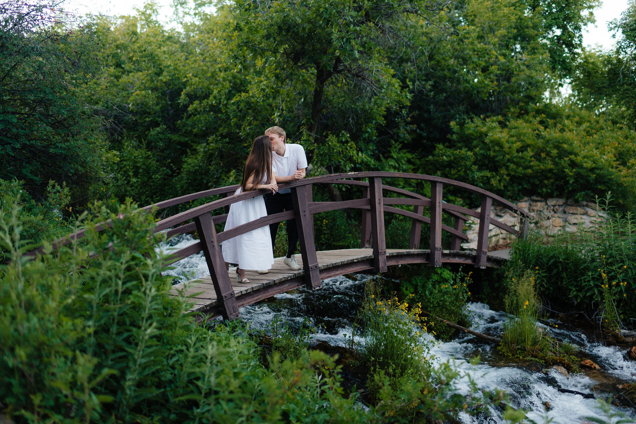 a couple kisses on a bridge over a river during their summer engagment photos in the alpine loop at american fork canyon and provo canyon, photographed by utah county engagement photographer sarah mismash