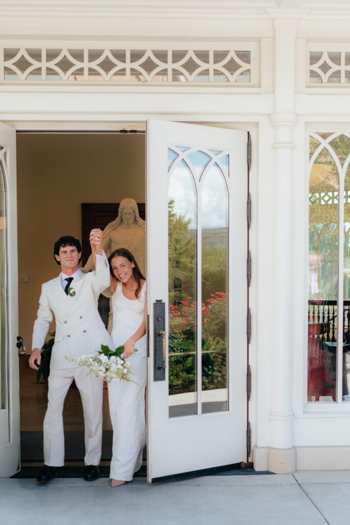 The couple exits through the temple's front doors, hands raised, celebrating their September wedding at the Provo City Center Temple.
