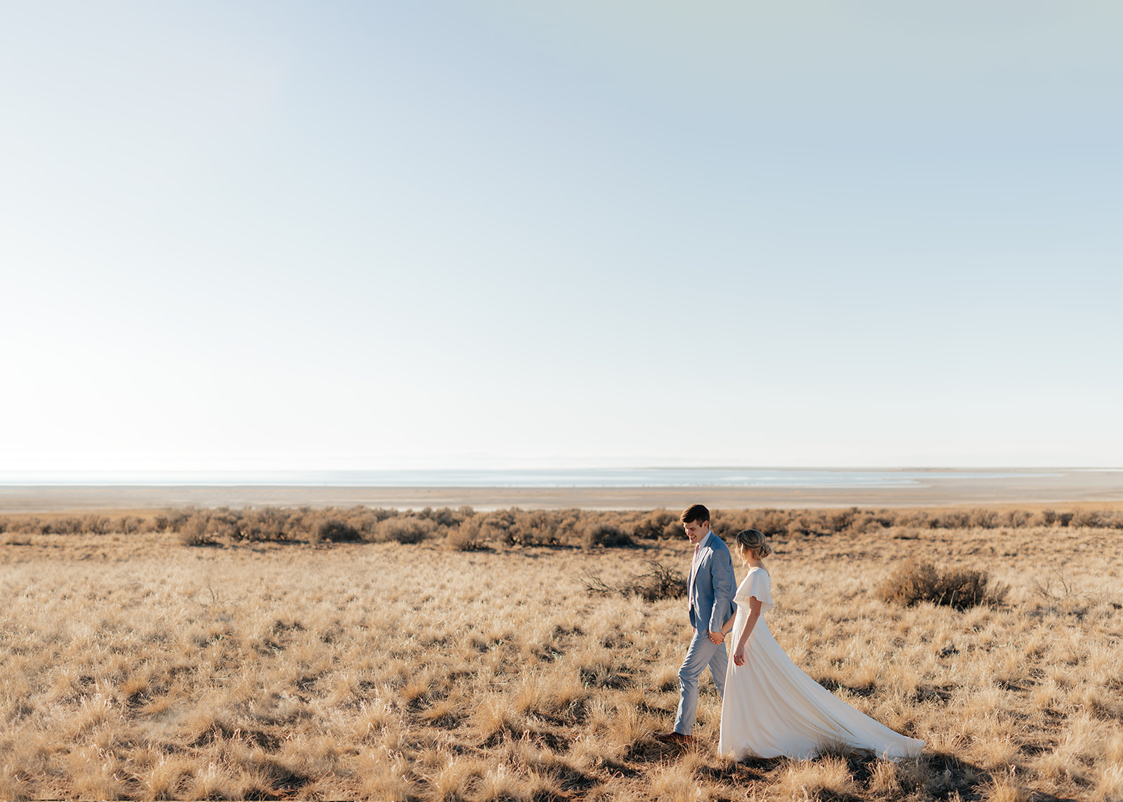 a couple walks together through the snow covered grass at antelope island in northern utah during a winter wedding portrait session with utah snowy winter photographer sarah mismash